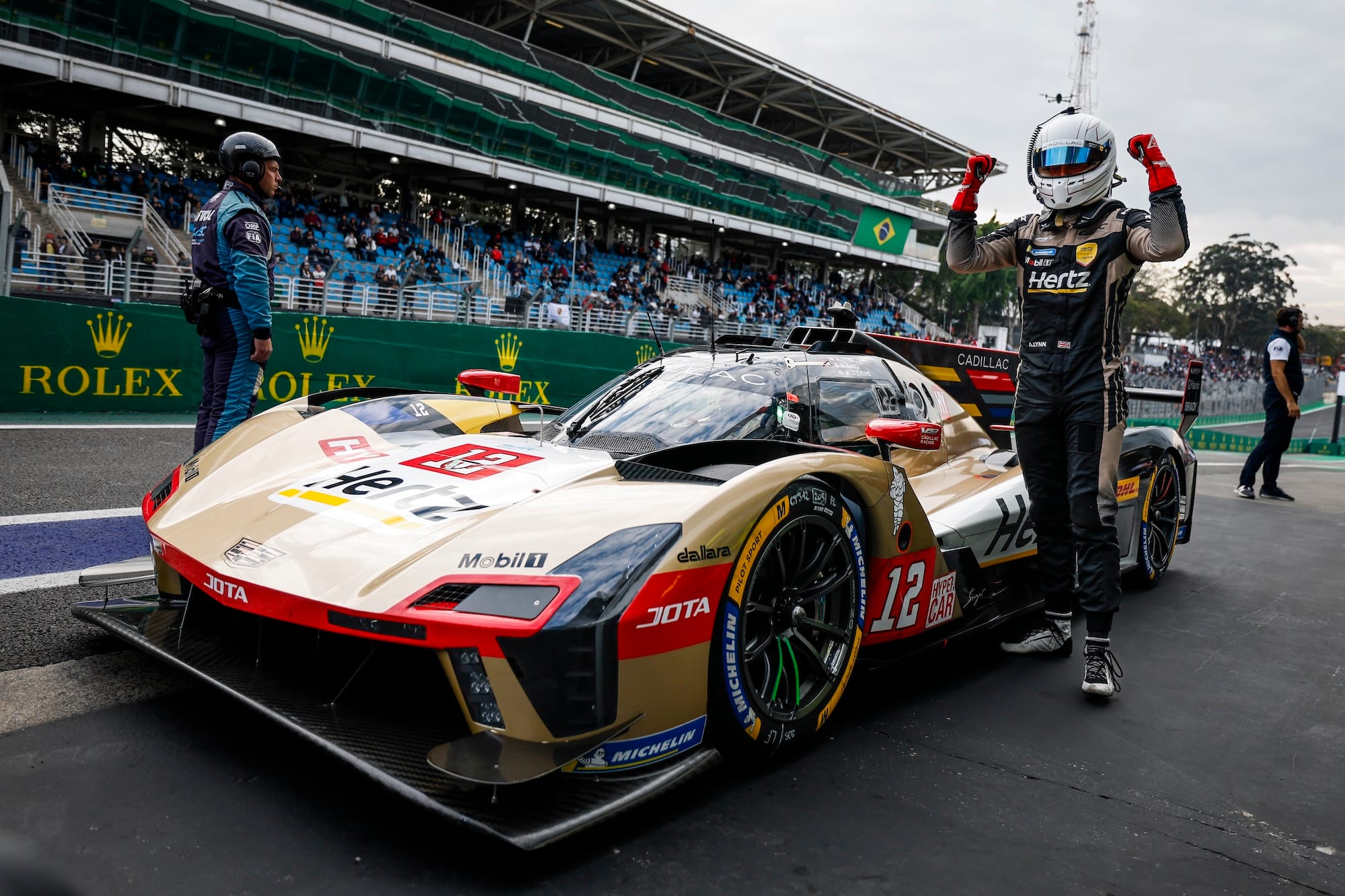 Alex Lynn standing next to his #12 Cadillac, with his helmet on and his arms raised, celebrating his pole position at Interlagos